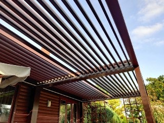 Zadaszenie tarasu z metalowych profili w kolorze ciemnego brązu, tworzące regularny wzór linii na tle błękitnego nieba. Widoczny fragment budynku z drewnianą elewacją i okno. Ujęcie z dołu...