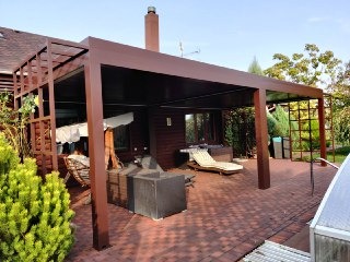 Zadaszony taras z brązową konstrukcją szkieletową, wyłożony czerwoną kostką brukową, z widocznymi meblami ogrodowymi i elementami zieleni w tle.