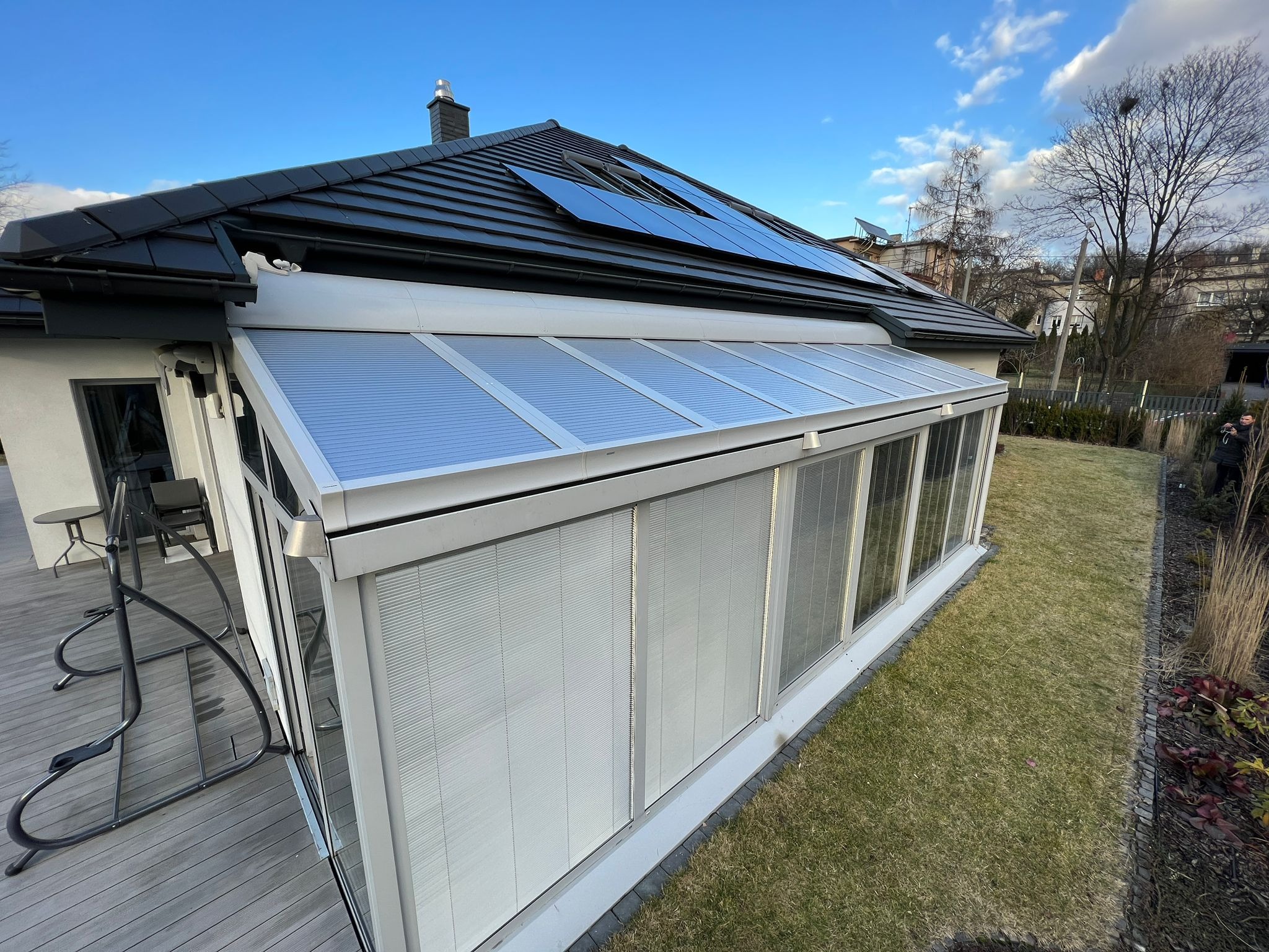 Zewnętrzny widok przeszklonej oranżerii z żaluzjami, dobudowanej do domu z czarnym dachem i panelami słonecznymi, w tle zielony trawnik i zabudowa miejska.