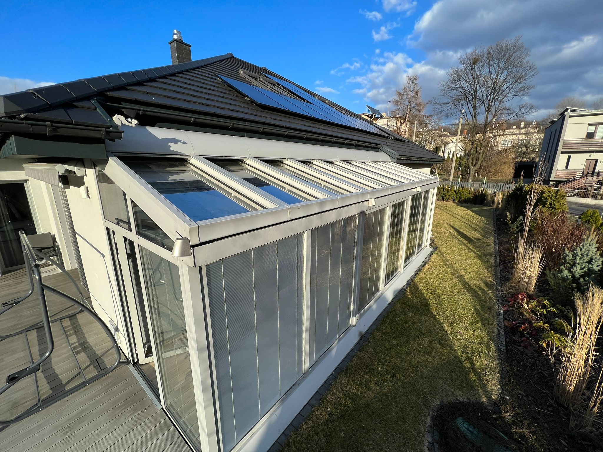 Nowoczesna szklana oranżeria z aluminiową konstrukcją przylegająca do domu z panelami słonecznymi na dachu, widok z zewnątrz w słoneczny dzień.