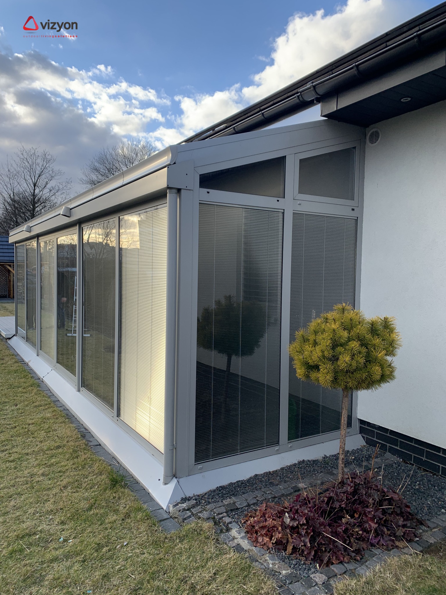 Szklana zabudowa tarasu z żaluzjami wewnętrznymi, przylegająca do białego budynku, z małym drzewkiem ozdobnym zasadzonym obok.