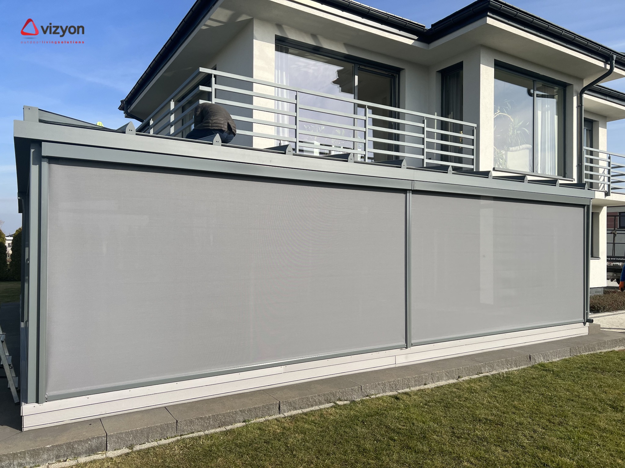 Zewnętrzne rolety przeciwsłoneczne w kolorze szarym, zamontowane na tarasie nowoczesnego domu z białymi ścianami i szarą balustradą. Widoczny fragment trawnika i błękitne niebo.