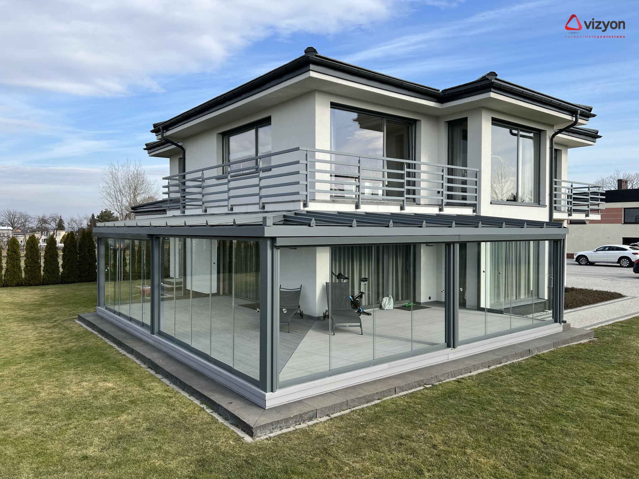 Nowoczesna przeszklona weranda z szarymi ramami, przylegająca do białego domu z balkonem z szarą balustradą, widok z zewnątrz na trawnik i błękitne niebo.