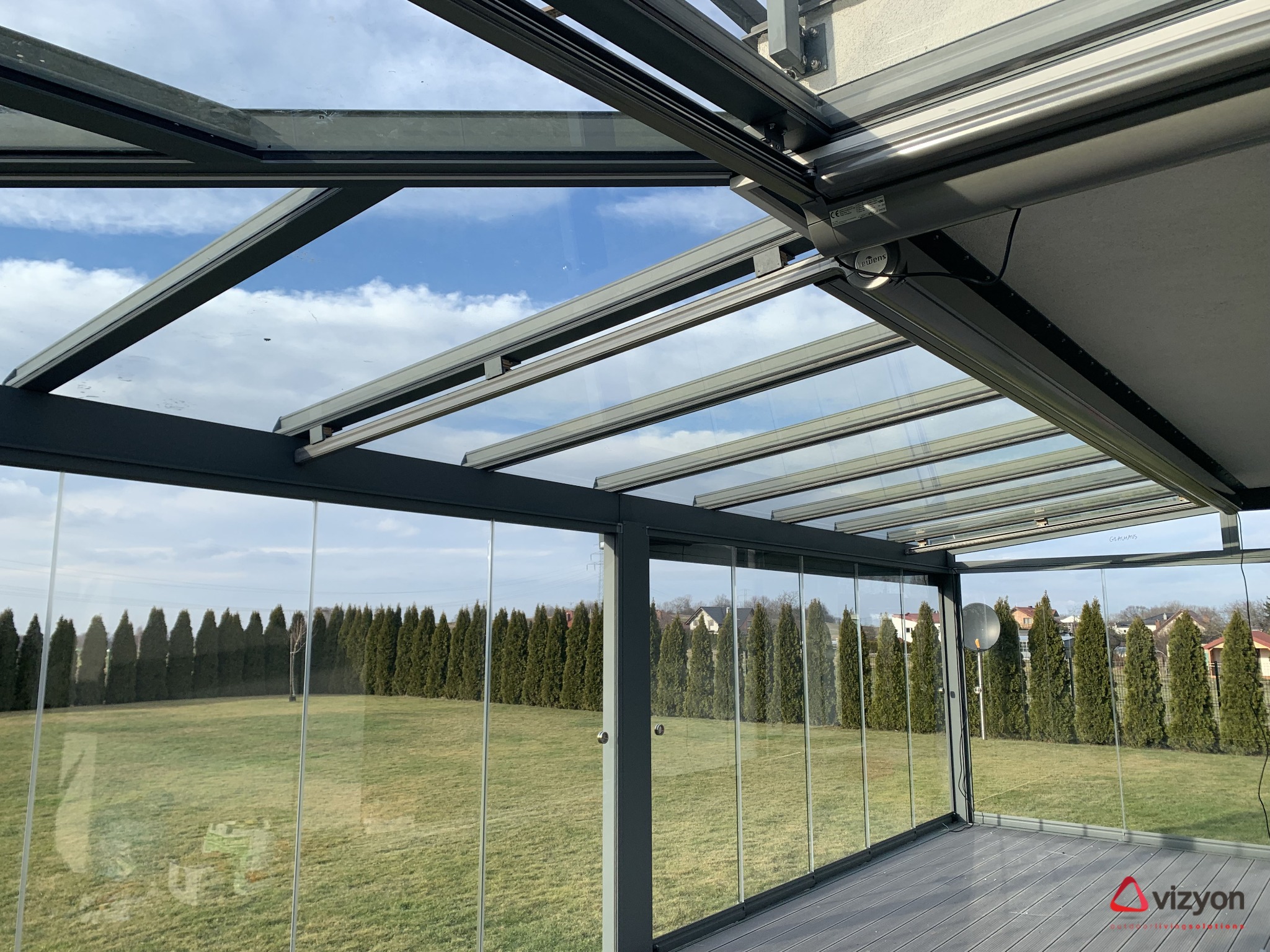 Szklana zabudowa tarasu z przesuwnymi drzwiami i zadaszeniem, widok z wnętrza na ogród z rzędem tuj i błękitne niebo z chmurami.