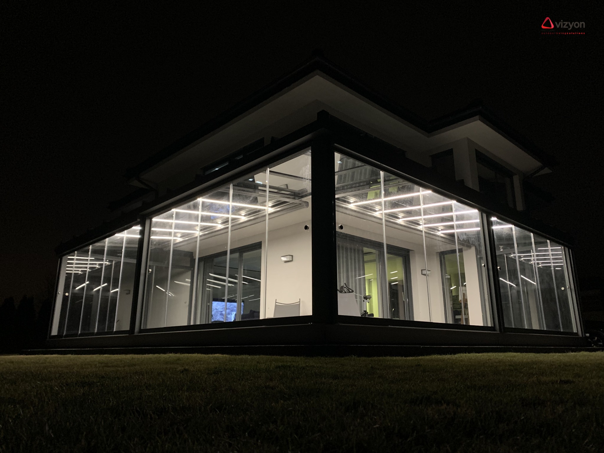 Nowoczesna, przeszklona oranżeria z balustradami szklanymi, oświetlona od wewnątrz, widoczna nocą z ogrodu.