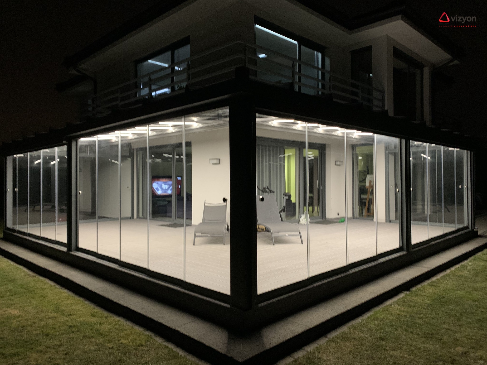 Nowoczesna szklana zabudowa tarasu z przesuwnymi drzwiami, oświetlona od wewnątrz, widok z zewnątrz w nocy, z widocznym fragmentem ogrodu i logo firmy w prawym górnym rogu.