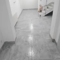 Wykończone wnętrze z szarymi płytkami podłogowymi, widoczne schody z metalową barierką i materiały budowlane w tle, ściany przygotowane do malowania.