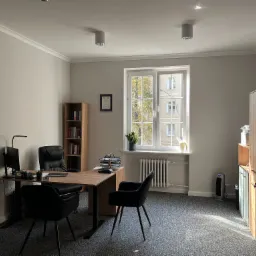 Jasne biuro z drewnianym biurkiem, czarnymi krzesłami i regałem z książkami, widok na drzewa przez okno, na ścianie certyfikat.