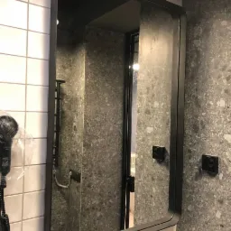 Łazienka z lustrem w czarnej ramie, białe płytki na ścianie po lewej stronie i szara ściana z mozaiką po prawej, widoczny suszarka do włos&oacute;w i czarna armatura.