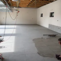 Układanie jasnych, kwadratowych płytek podłogowych w garażu, widoczna kałuża wody i glazurnik wycierający podłogę, ściany przygotowane do malowania, surowy strop z widocznymi belkami.