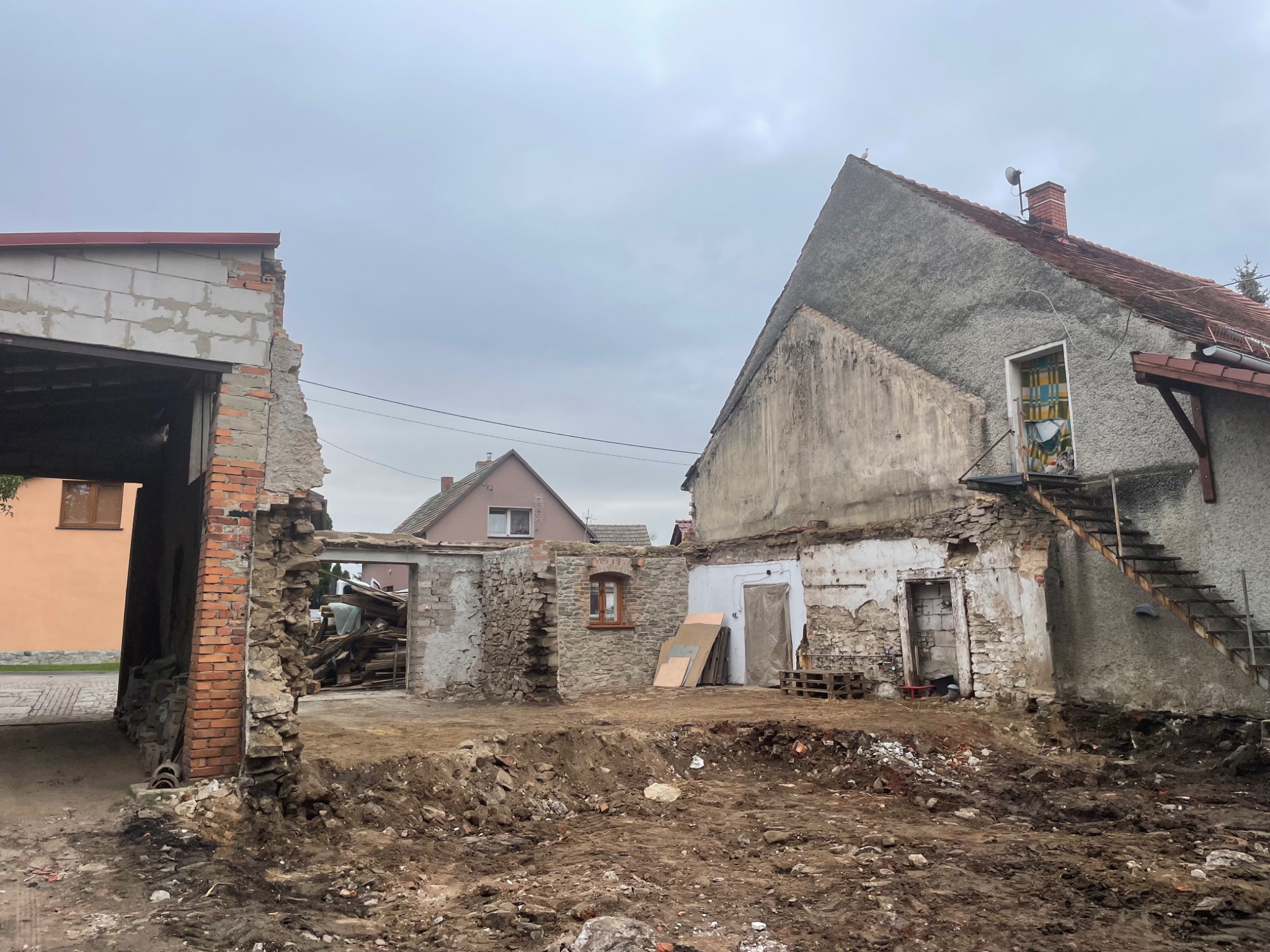 Częściowo zburzony budynek mieszkalny z odsłoniętymi cegłami i gruzem, widoczne są pozostałości ścian i fundamentów, a w tle inne budynki mieszkalne pod pochmurnym niebem.