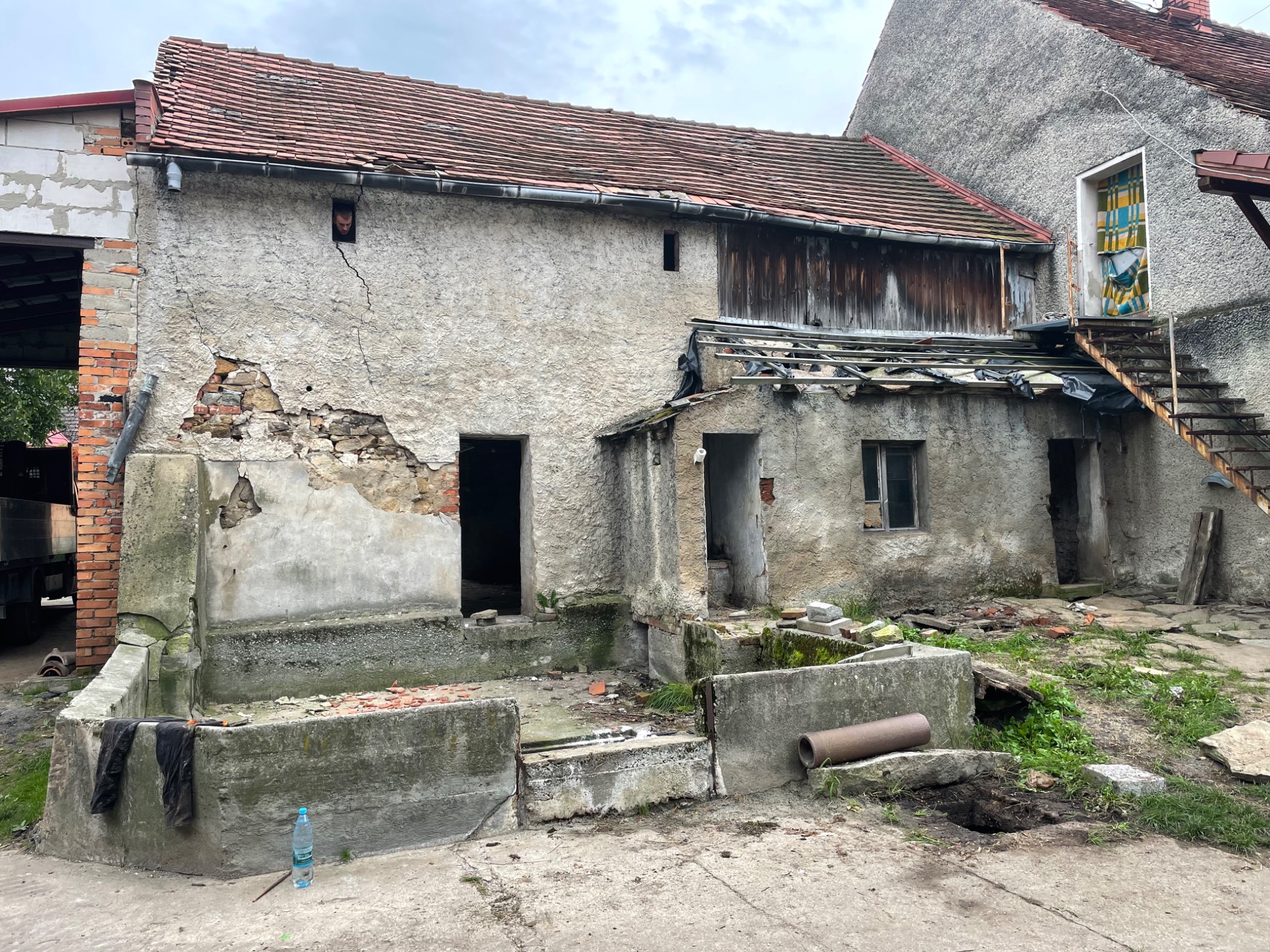 Stary, zaniedbany budynek gospodarczy z częściowo zawalonymi ścianami, widoczne uszkodzenia muru, odkryta cegła, rdzewiejące schody zewnętrzne, butelka wody i robocze spodnie leżące na betonowej...