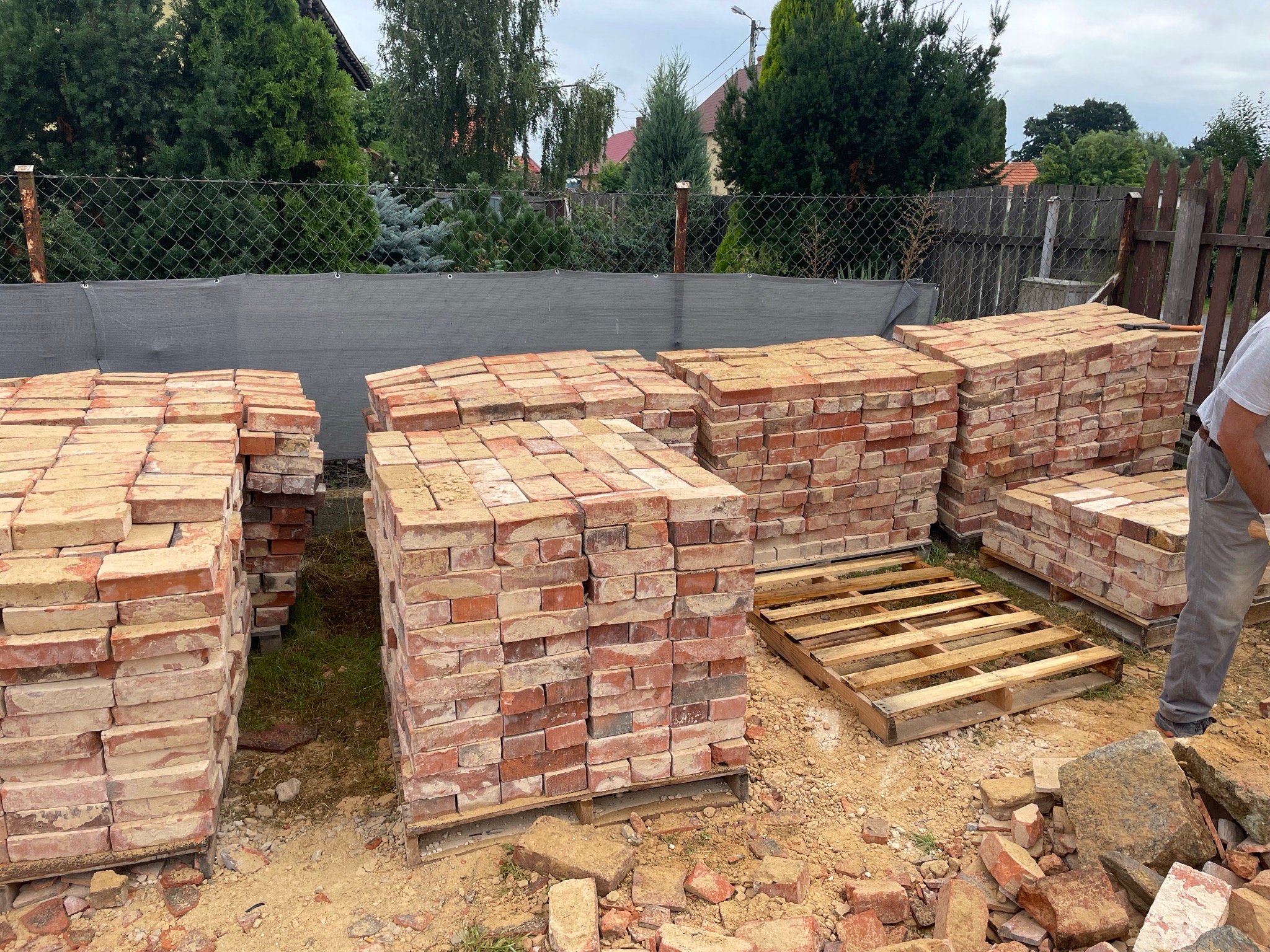 Stosy czerwonej cegły na paletach na placu budowy, widoczny fragment ogrodzenia z siatki i zieleń w tle, obok cegieł leżą gruz i resztki cegieł.