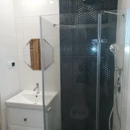 Nowoczesna łazienka z kabiną prysznicową walk-in, czarnymi płytkami o trójwymiarowej fakturze, białą umywalką z szafką i lustrem w drewnianej ramie.