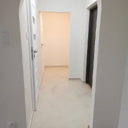 Wnętrze korytarza po remoncie z łukowym przejściem, białymi ścianami, jasną podłogą z płytek imitujących marmur oraz ciemną, wbudowaną szafą w głębi. Widoczne dwie białe, wewnętrzne drzwi...