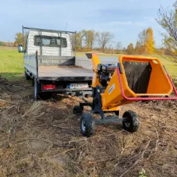 Pomarańczowy rębak do gałęzi Master z silnikiem Loncin, podłączony do białego samochodu dostawczego z otwartą skrzynią ładunkową, na polu z gałęziami i ściętą trawą.