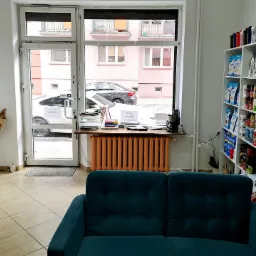 Wnętrze gabinetu z regałami pełnymi suplementów diety i odżywek, widok z poczekalni z turkusową sofą, okno z widokiem na ulicę, w tle grzejnik pomalowany na pomarańczowo.