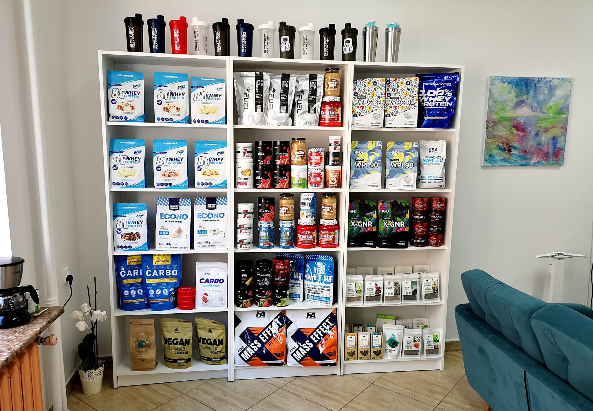Półka z suplementami diety i odżywkami dla sportowców, różnorodne opakowania białek, kreatyny, BCAA i innych preparatów, obok sofa w kolorze turkusowym i obraz na ścianie.