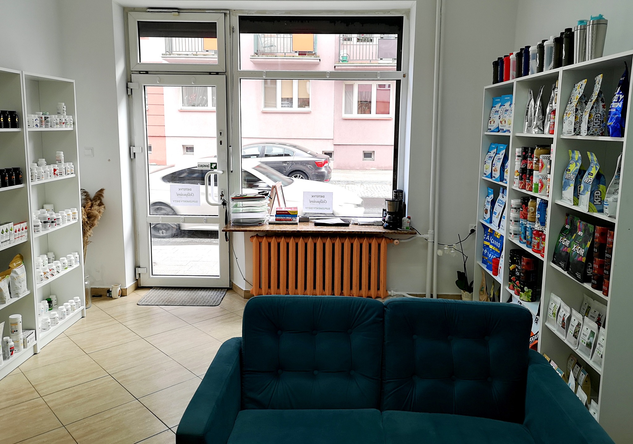 Wnętrze gabinetu z regałami pełnymi suplementów diety i odżywek, widok z poczekalni z turkusową sofą, okno z widokiem na ulicę, w tle grzejnik pomalowany na pomarańczowo.