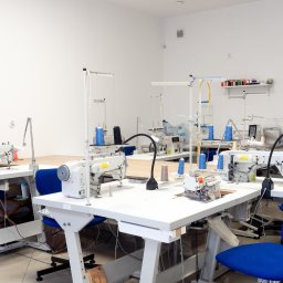 JUST LOOK - Jasne, przestronne wnętrze szwalni z kilkoma stanowiskami pracy wyposażonymi w białe stoły i maszyny do szycia. Na półce widoczne kolorowe nici.