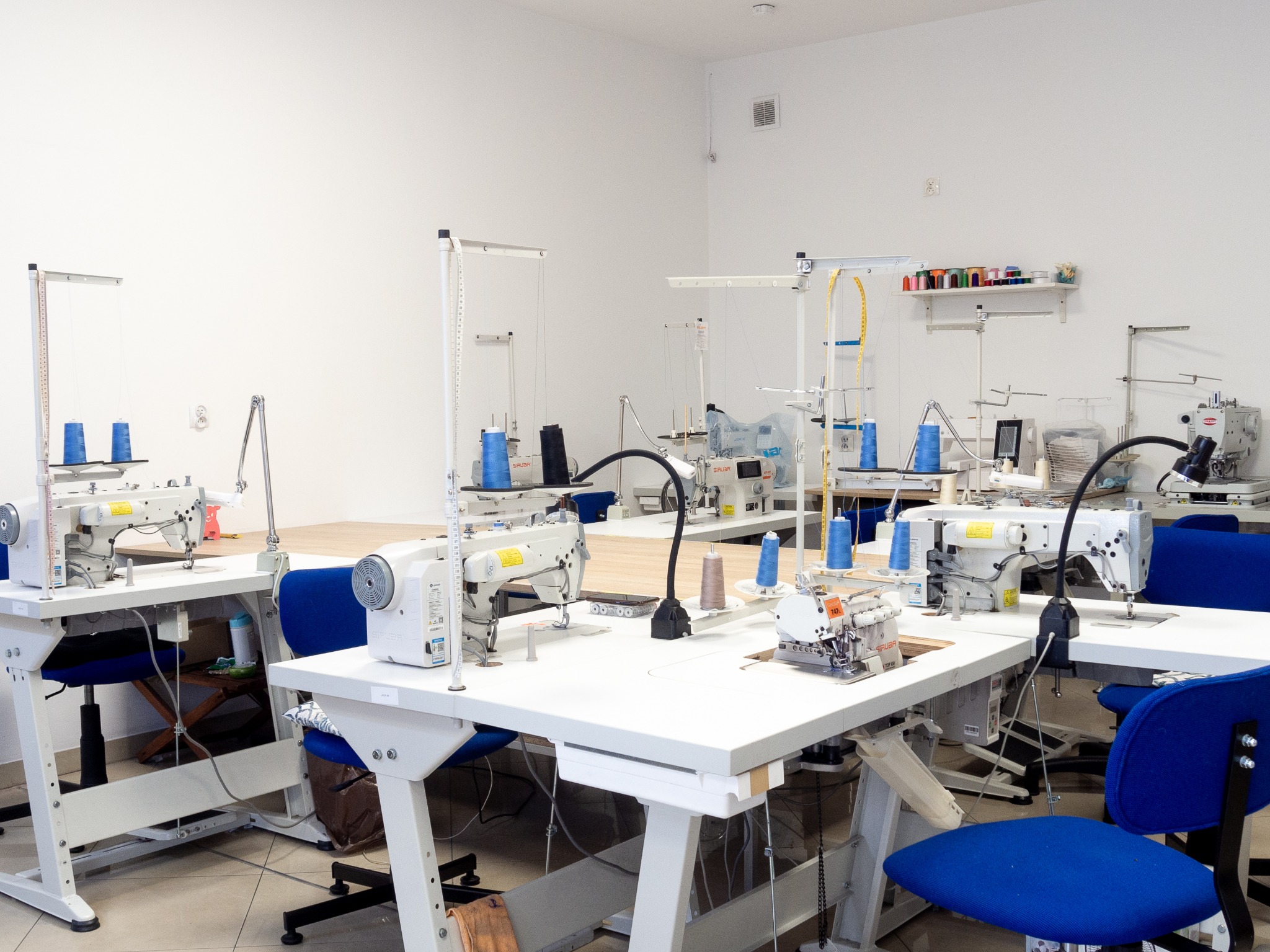 Jasne, przestronne wnętrze szwalni z kilkoma stanowiskami pracy wyposażonymi w białe stoły i maszyny do szycia. Na półce widoczne kolorowe nici.