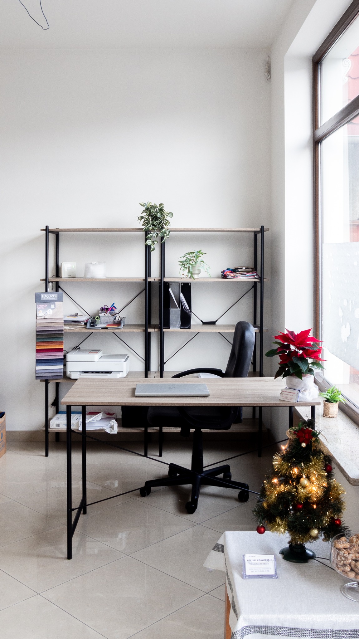 Jasne biuro krawieckie z wzornikiem tkanin, biurkiem z laptopem, regałem i dekoracją świąteczną przy oknie. Minimalistyczny wystrój z roślinami.