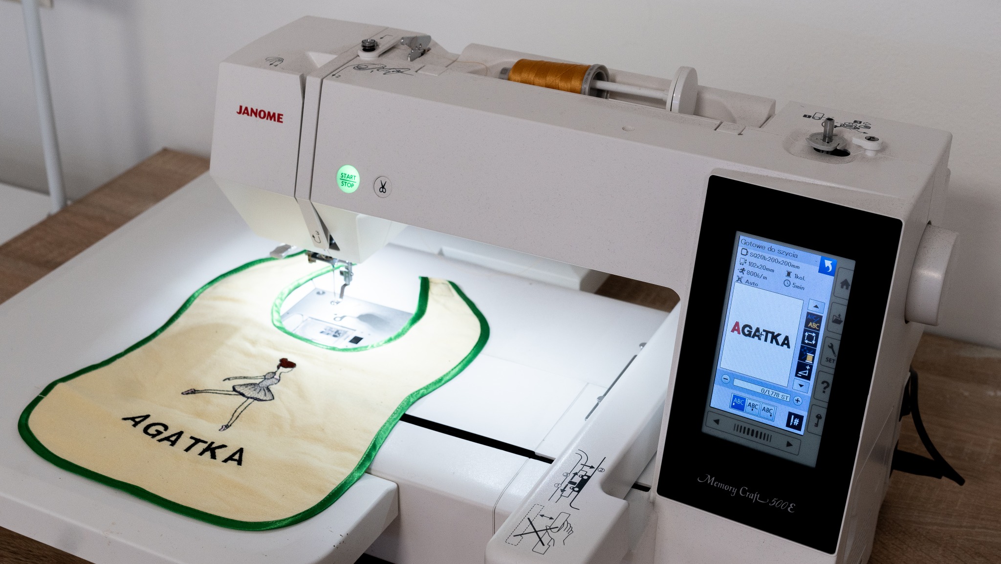 Biała maszyna Janome haftuje imię 'Agatka' i baletnicę na beżowym śliniaku z zieloną lamówką. Ekran maszyny pokazuje wzór do haftu.