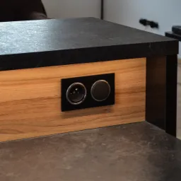 Fragment blatu z czarnego kamienia i drewnianej obudowy z gniazdkiem elektrycznym i włącznikiem światła w czarnej ramce.