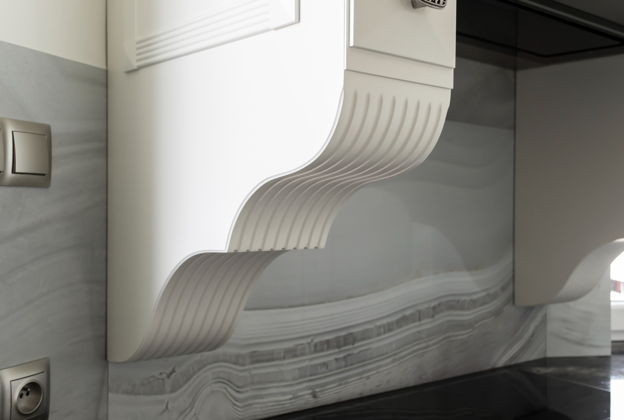 Detale wykończenia białej szafki kuchennej z dekoracyjnym, frezowanym elementem podtrzymującym, na tle marmurowej ściany i blatu.