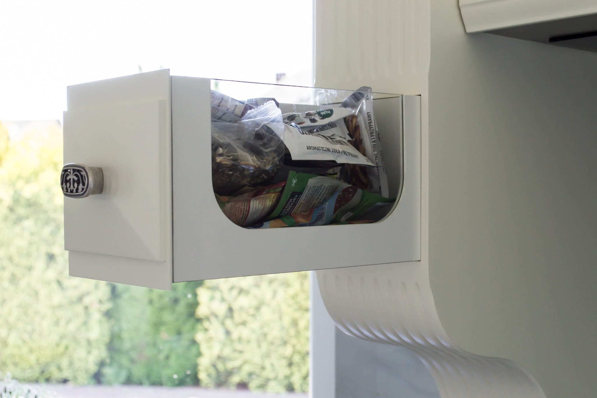 Otwarta, biała szafka kuchenna z przezroczystym frontem, wypełniona torebkami z przyprawami, widoczna na tle okna z zielenią. Dekoracyjny uchwyt szafki.