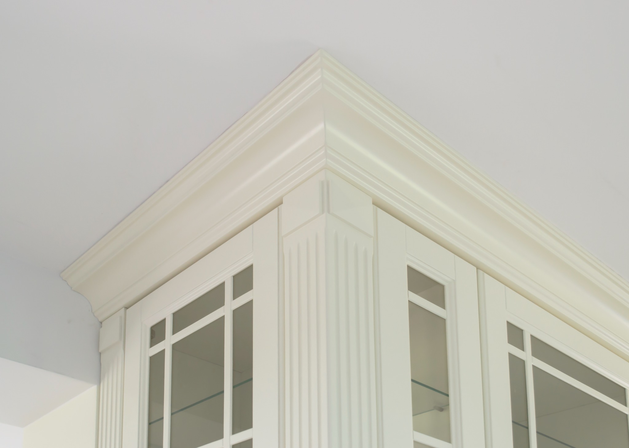 Detale wykończenia narożnego białej szafki kuchennej z przeszklonymi drzwiczkami i dekoracyjnym gzymsem.