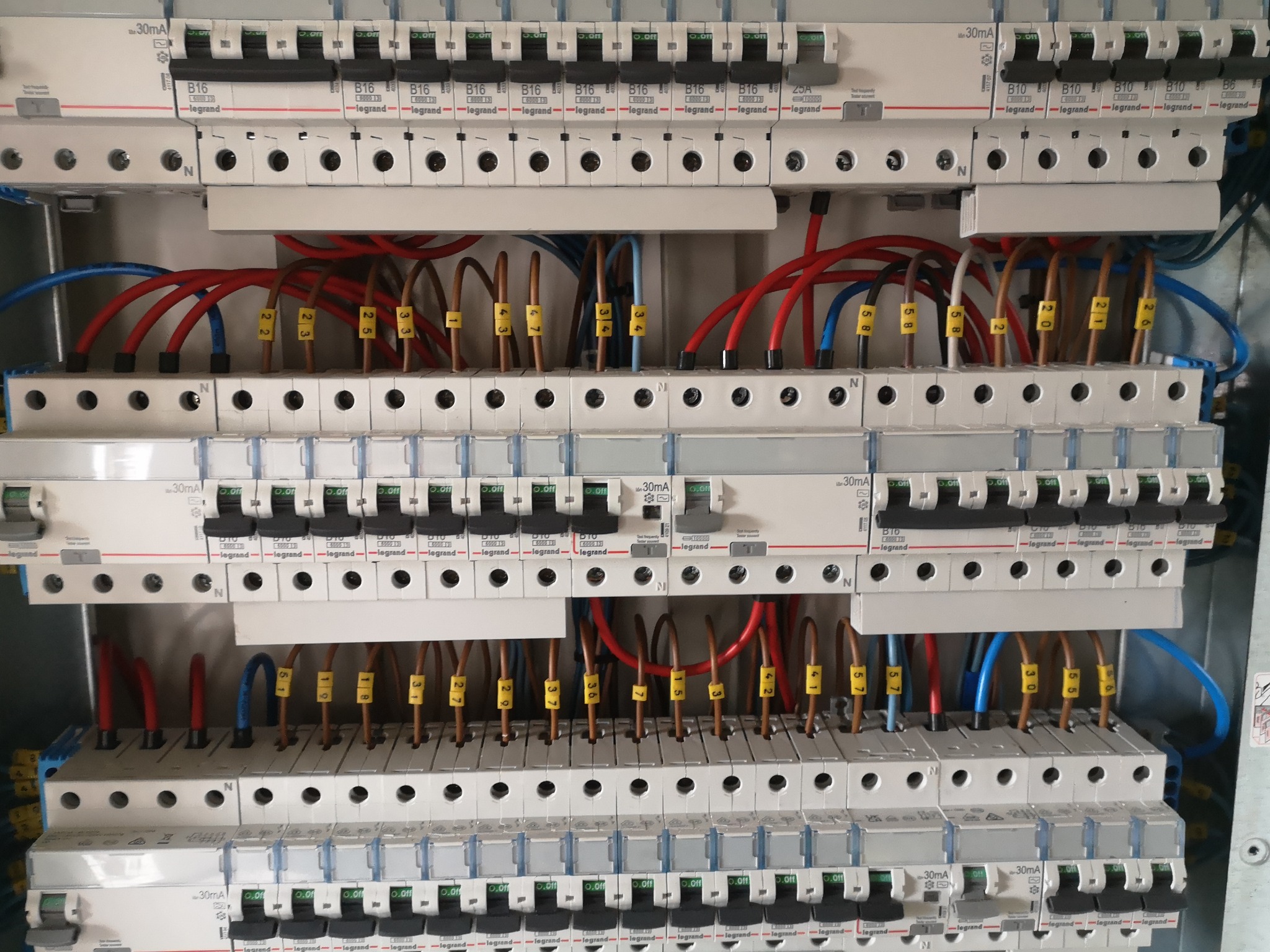 Rozdzielnica elektryczna z widocznymi bezpiecznikami, przewodami oznaczonymi numerowanymi tulejkami i elementami marki Legrand.