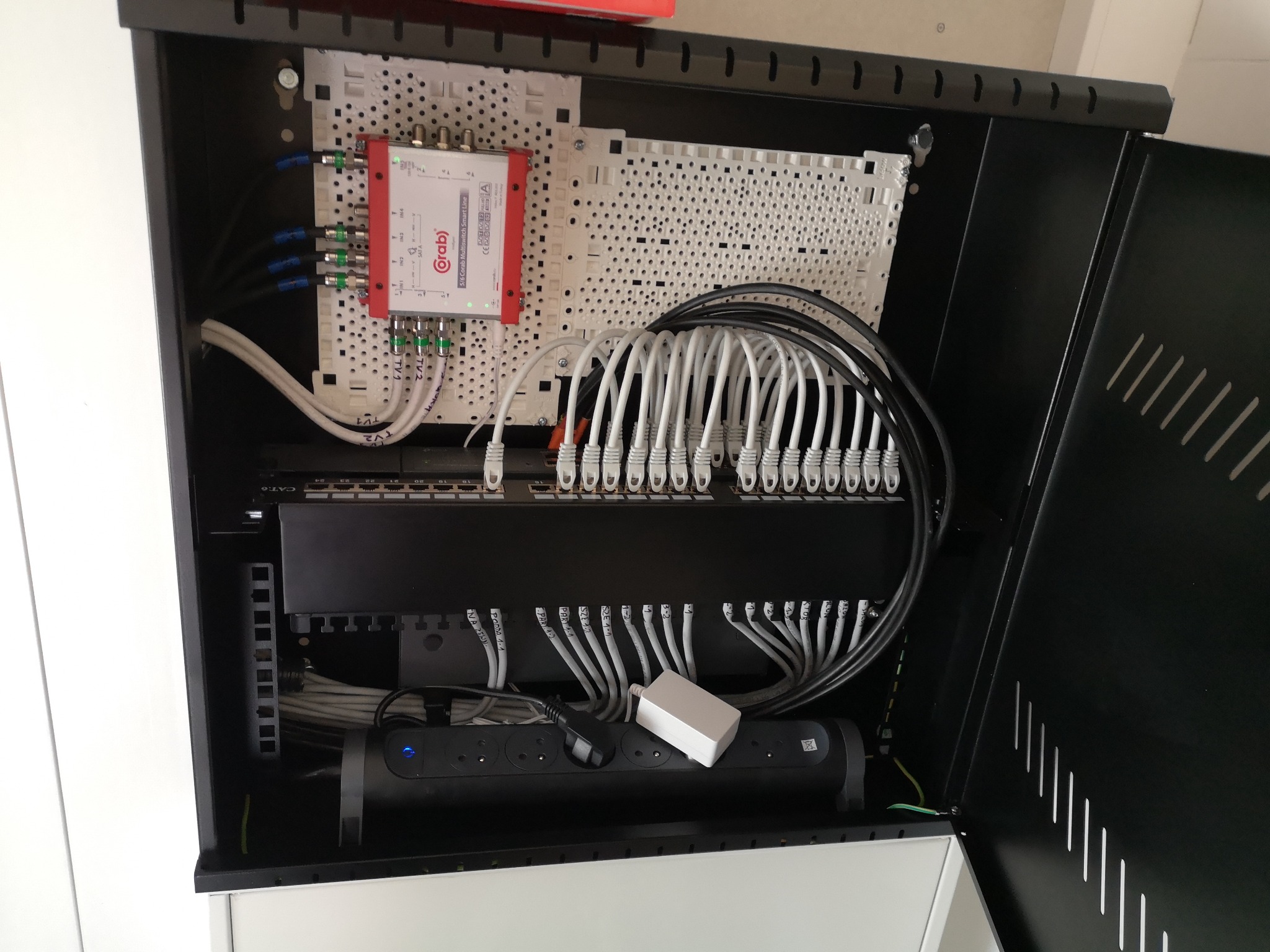 Otwarta szafa rack z zamontowanymi elementami sieci telekomunikacyjnej: multiswitch Corab, patch panel z podłączonymi kablami Ethernet, listwa zasilająca z wtyczką i zasilaczem. Widoczne etykiety...