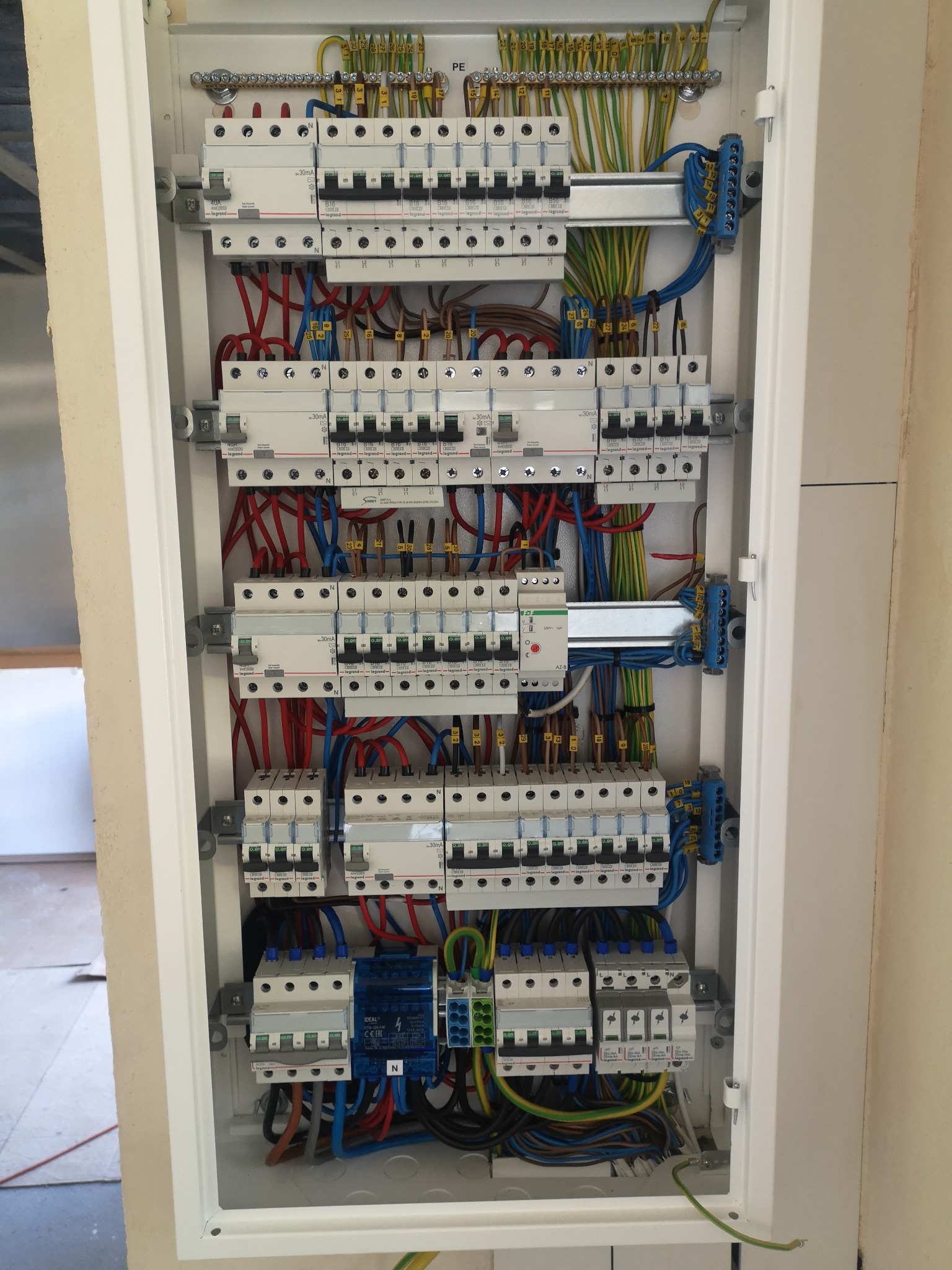 Otwarta skrzynka elektryczna z zainstalowanymi bezpiecznikami, przewodami w kolorach czerwonym, niebieskim, zielono-żółtym i brązowym, widoczne elementy montażowe i oznaczenia na urządzeniach.