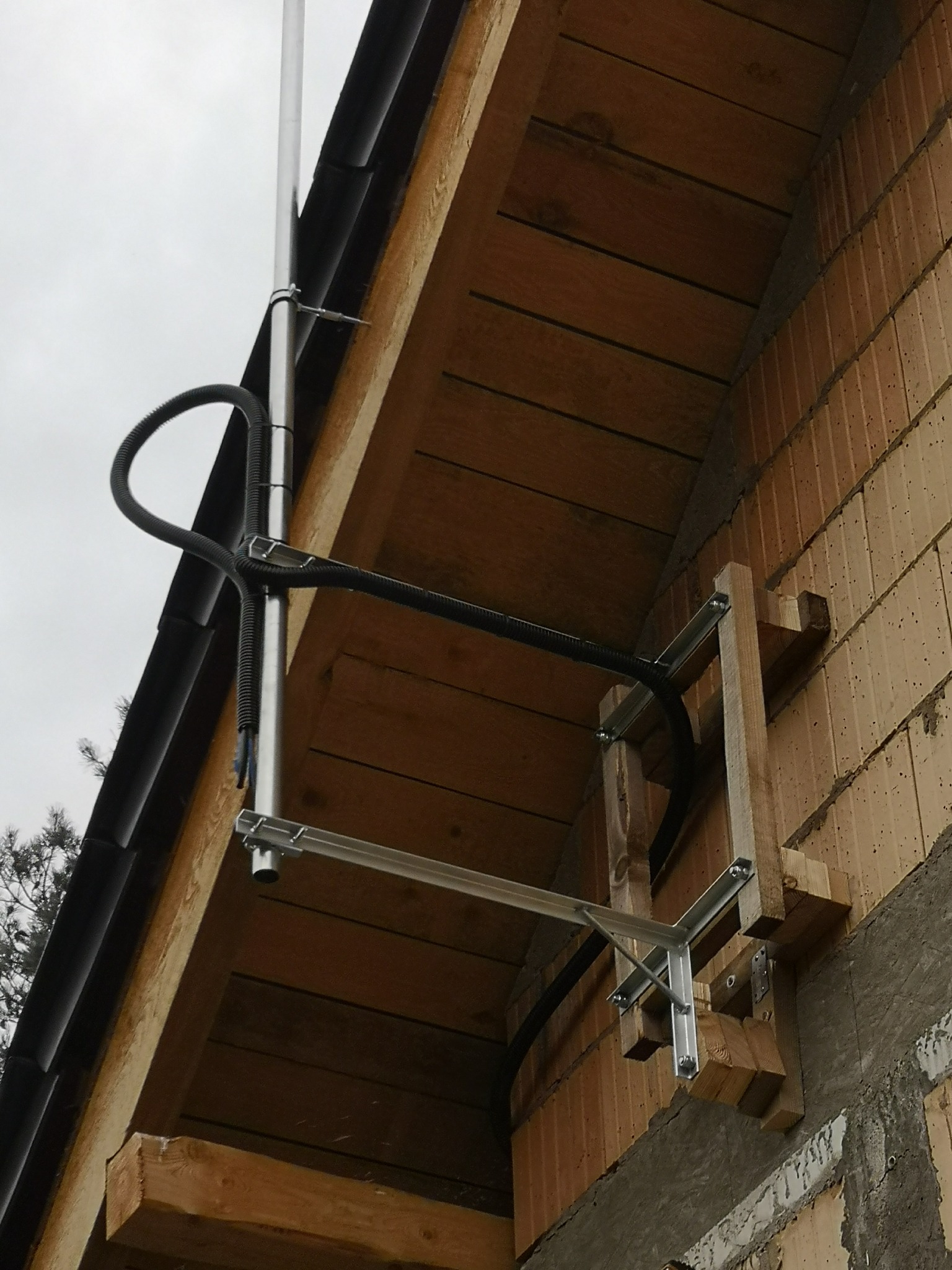 Metalowa instalacja odgromowa przymocowana do ściany budynku z cegły, z widocznymi elementami drewnianymi i czarną rurką ochronną kabla.