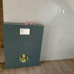 Stelaż podtynkowy WC w trakcie montażu, widoczne przyłącza kanalizacyjne i fragment ściany wyłożonej płytkami imitującymi marmur.