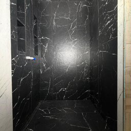 BUD VADYM SYLCHENKO - Kabina prysznicowa wyłożona czarnymi płytkami imitującymi marmur z białymi żyłami, z wnękami na kosmetyki. Widoczny odpływ liniowy i fragment ściany w trakcie wykończenia.