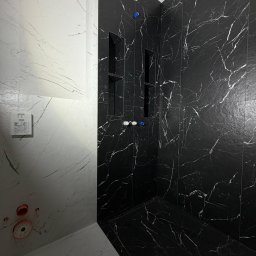 BUD VADYM SYLCHENKO - Wykończona łazienka z czarno-białymi płytkami imitującymi marmur. Widoczne wnęki w ścianie wyłożonej czarnymi płytkami i elementy instalacji wodnej.