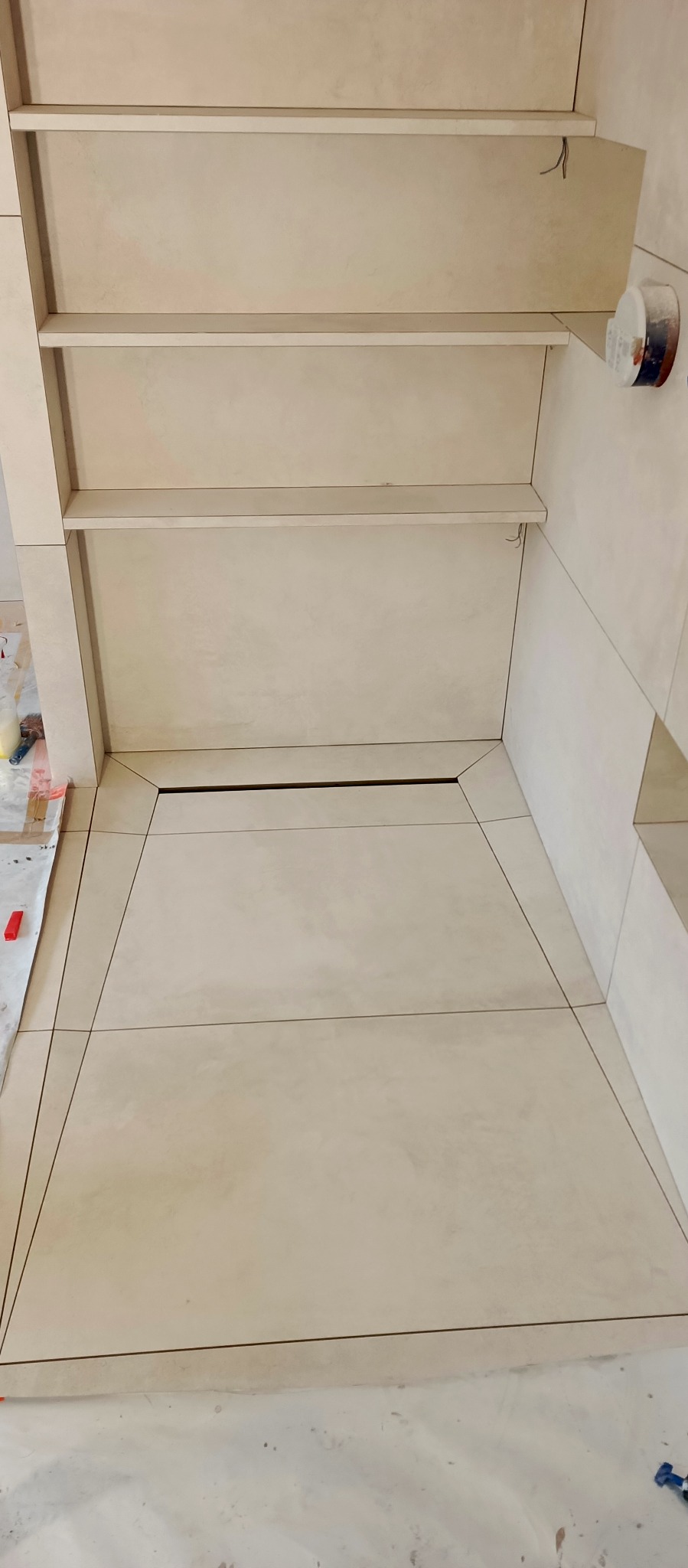 Beżowa łazienka w trakcie wykańczania, widok z góry ukazuje ułożone płytki podłogowe z ciemnymi fugami, zabudowane półki ścienne i zabezpieczone folią malarską podłoże.