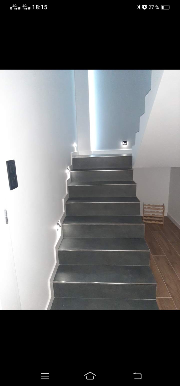 Nowoczesne schody z ciemnoszarej płytki z subtelnym oświetleniem LED pod każdym stopniem, białe ściany i drewniana podłoga w tle, widoczny fragment regału na wino.