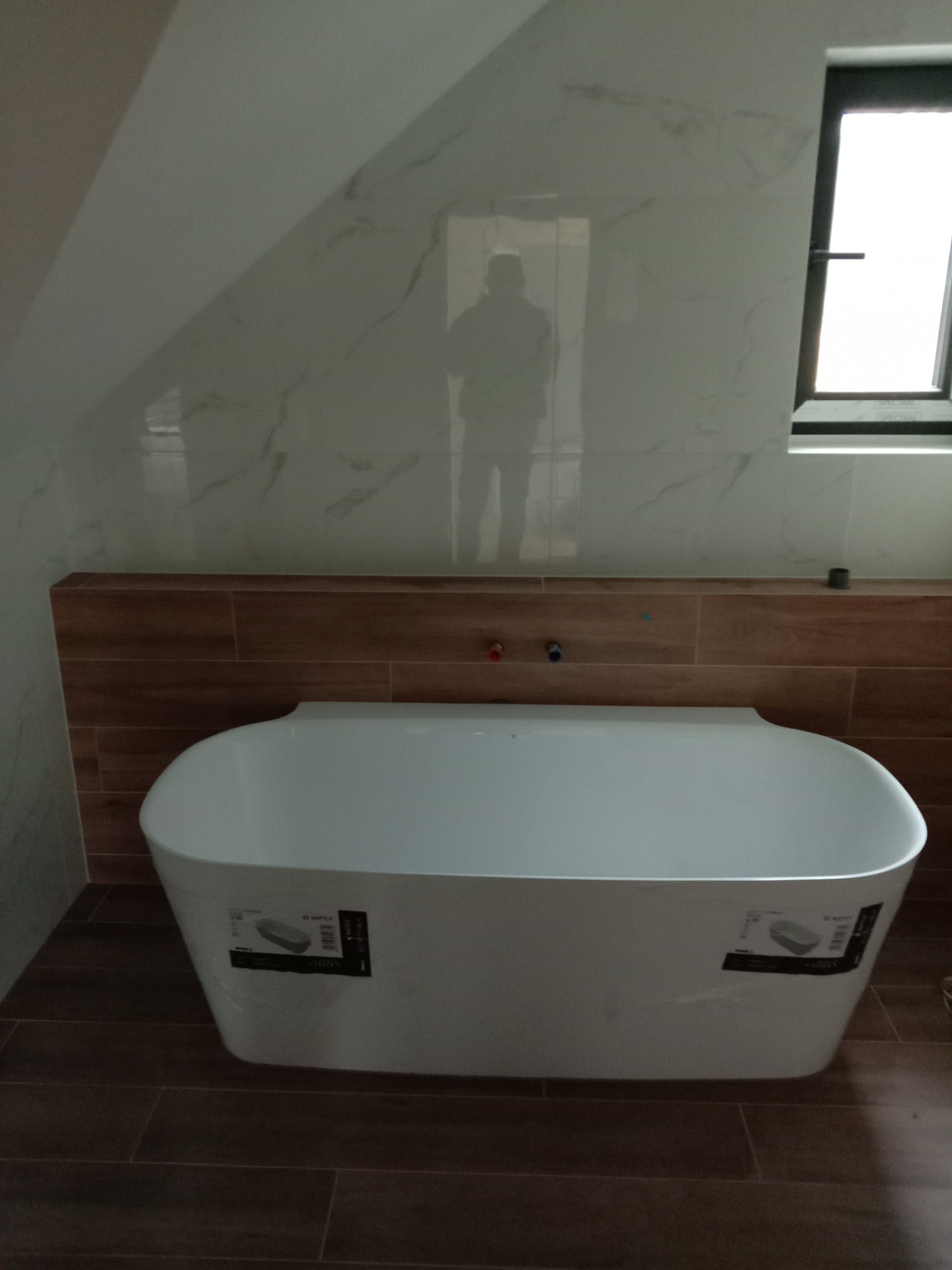 Nowoczesna wanna wolnostojąca w jasnej łazience z marmurowymi płytkami na ścianach i drewnopodobnymi płytkami za wanną i na podłodze. Widoczny sylwetkowy cień osoby na ścianie.