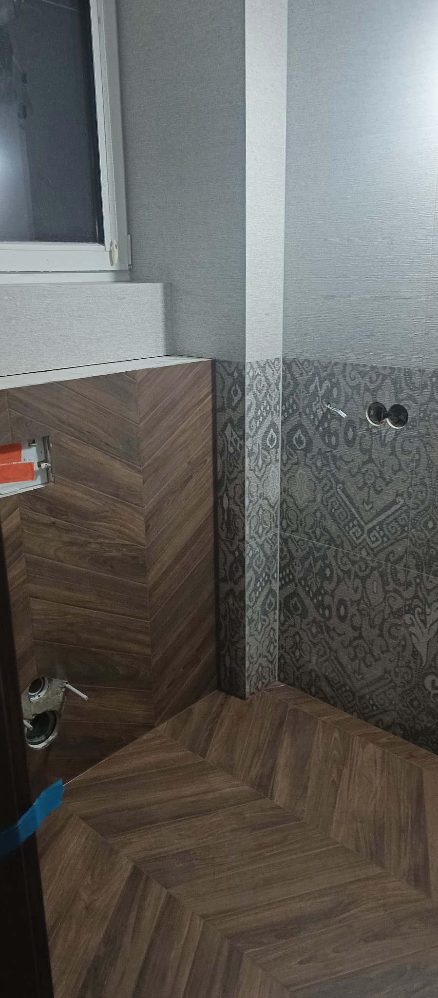 Wykończona łazienka z brązowymi płytkami ułożonymi w jodełkę na podłodze i ścianie, kontrastującymi z szarymi płytkami z wzorem na ścianie, widoczne gniazdka elektryczne bez osprzętu.