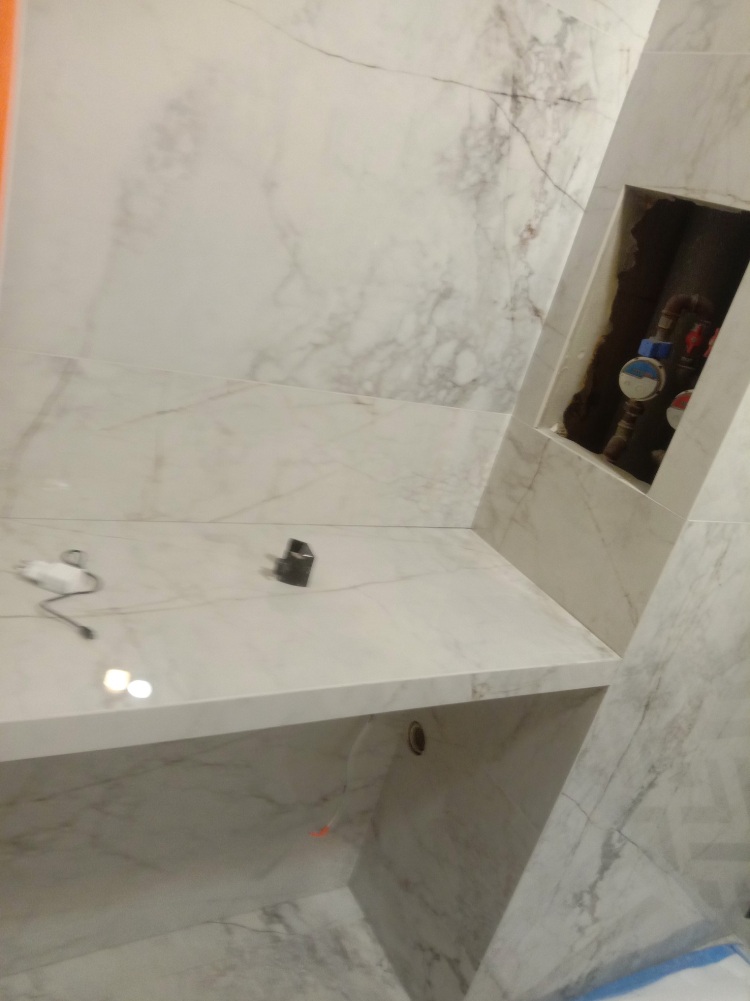 Wykończona wnęka z rurami i licznikami, obłożona białymi płytkami imitującymi marmur, z półką i ładowarką do telefonu na blacie