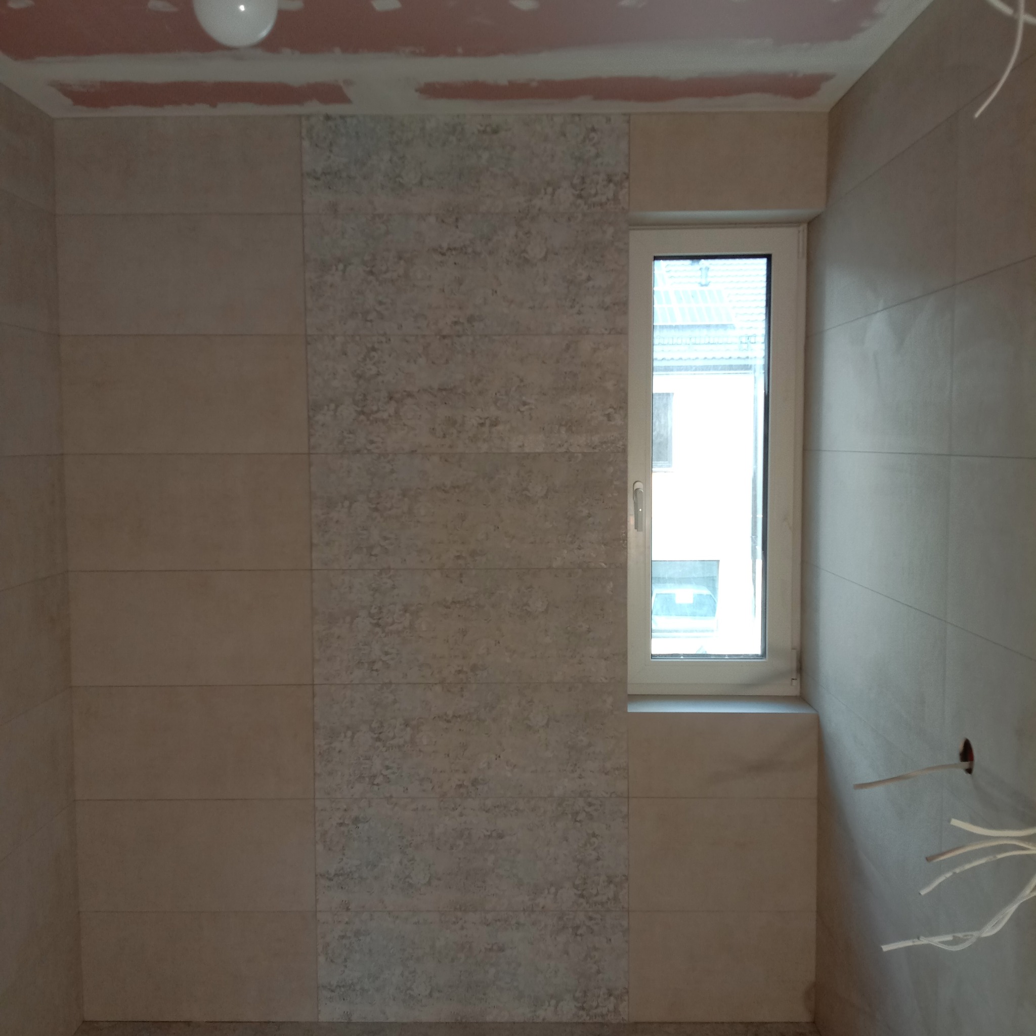 Ściana łazienki z jasnymi, poziomymi płytkami ceramicznymi o różnej fakturze, obok okna. Widoczne wystające przewody elektryczne i niedokończone wykończenie sufitu.