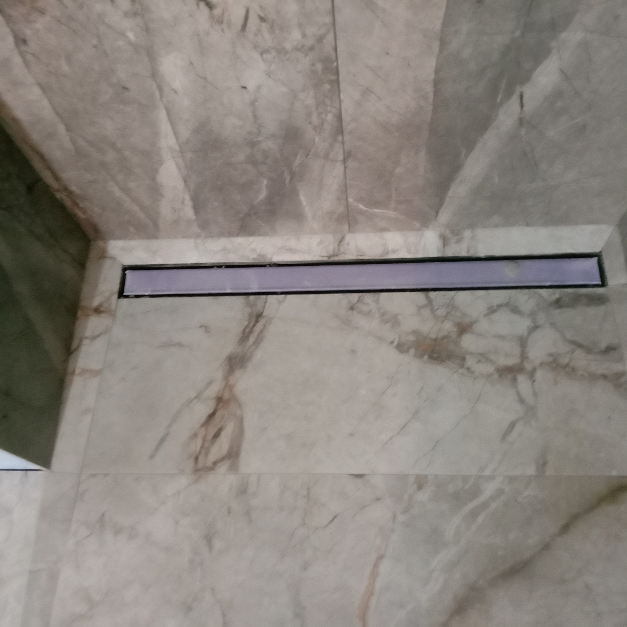 Wykończona podłoga prysznica z szarych płytek imitujących marmur i liniowym odpływem w rogu.