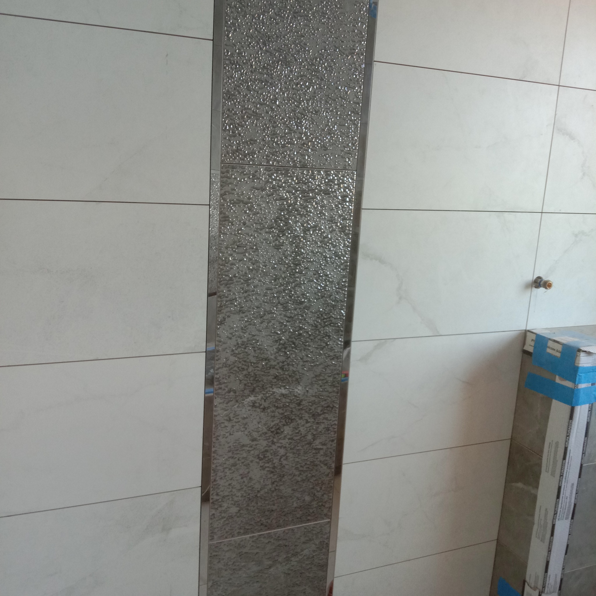 Wykończona ściana łazienki z białymi płytkami imitującymi marmur i dekoracyjnym pionowym pasem z mozaiki szklanej w srebrnej ramie.