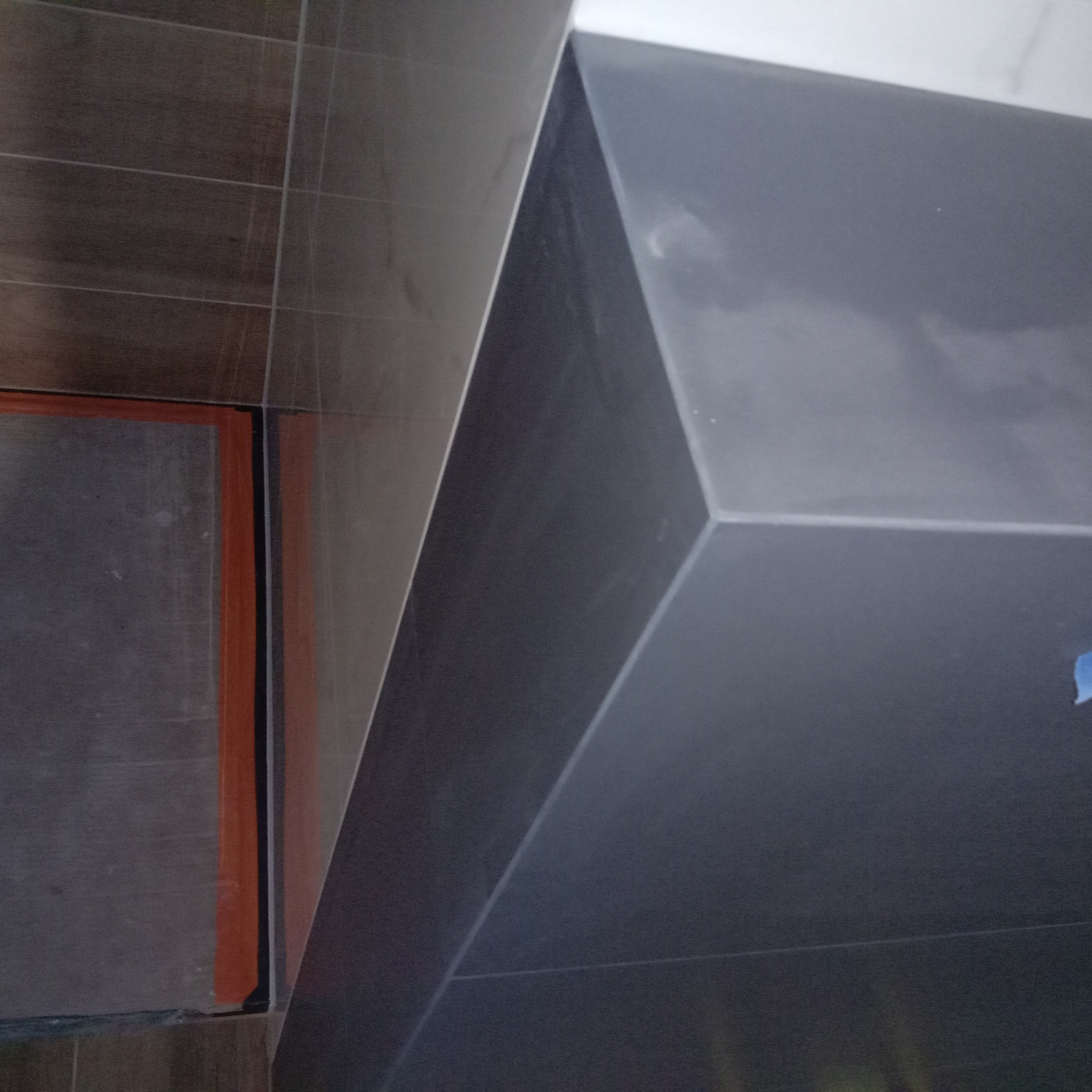 Ciemne, grafitowe płytki wielkoformatowe ułożone pod kątem na ścianie w łazience, widoczny fragment brązowej ściany z jasnymi fugami i pomarańczowym wykończeniem wokół otworu.