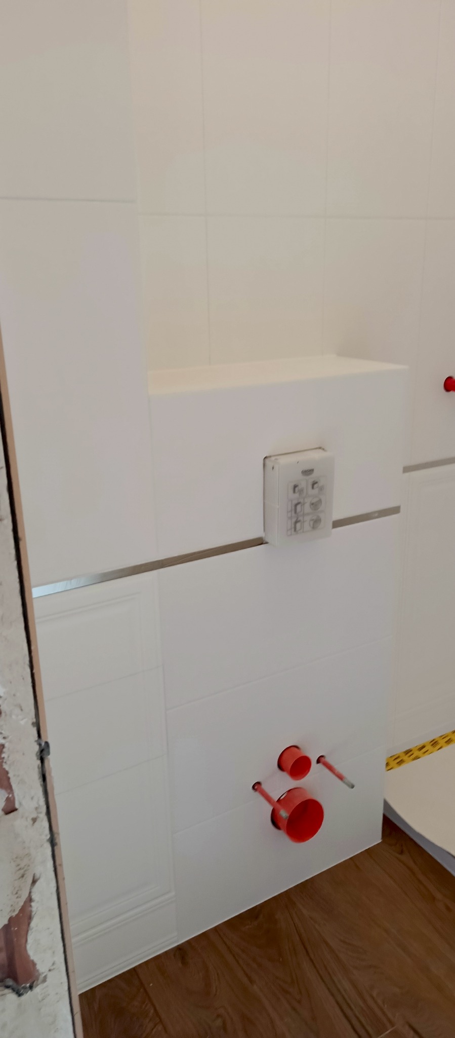 Białe płytki ścienne z instalacją podtynkową i przyciskiem spłukującym w łazience. Widoczne czerwone rury i drewnopodobna podłoga. Częściowo wykończone.