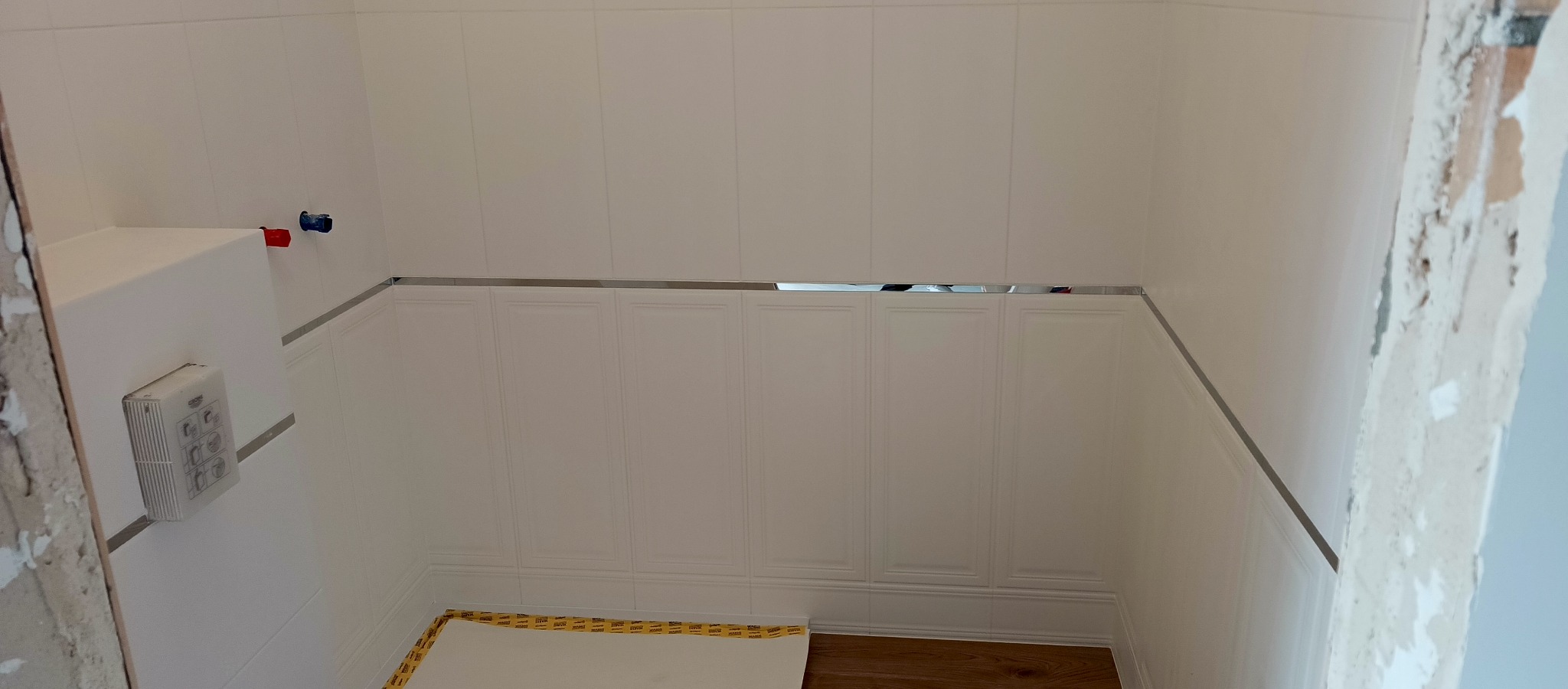 Białe panele ścienne z dekoracyjnymi listwami w łazience, widoczne przyłącza wodne i fragment podłogi z zabezpieczeniem. Ściana w trakcie prac wykończeniowych.