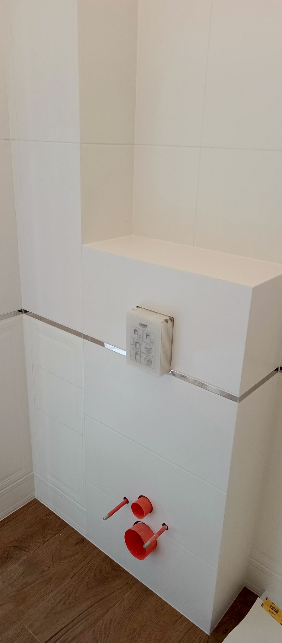 Biała zabudowa WC z widocznymi przyłączami wodno-kanalizacyjnymi i chromowanymi listwami wykończeniowymi. Podłoga z paneli imitujących drewno.